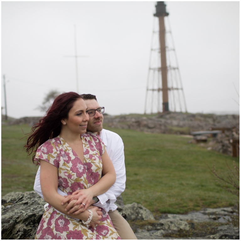 Engaged couple looks out to sea with Marblehead lighthouse in background
